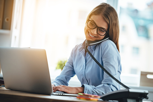 Cheerful young beautiful woman in glasses talking on the phone and using laptop with smile while sitting at her working place