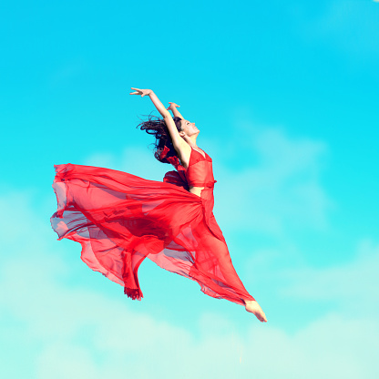 Girl in airy red dress jumping in the air, blue sky background
