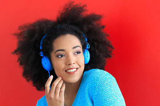 Cheerful young curly hair woman with blue headphones and blouse,  listening music. On red bacground.