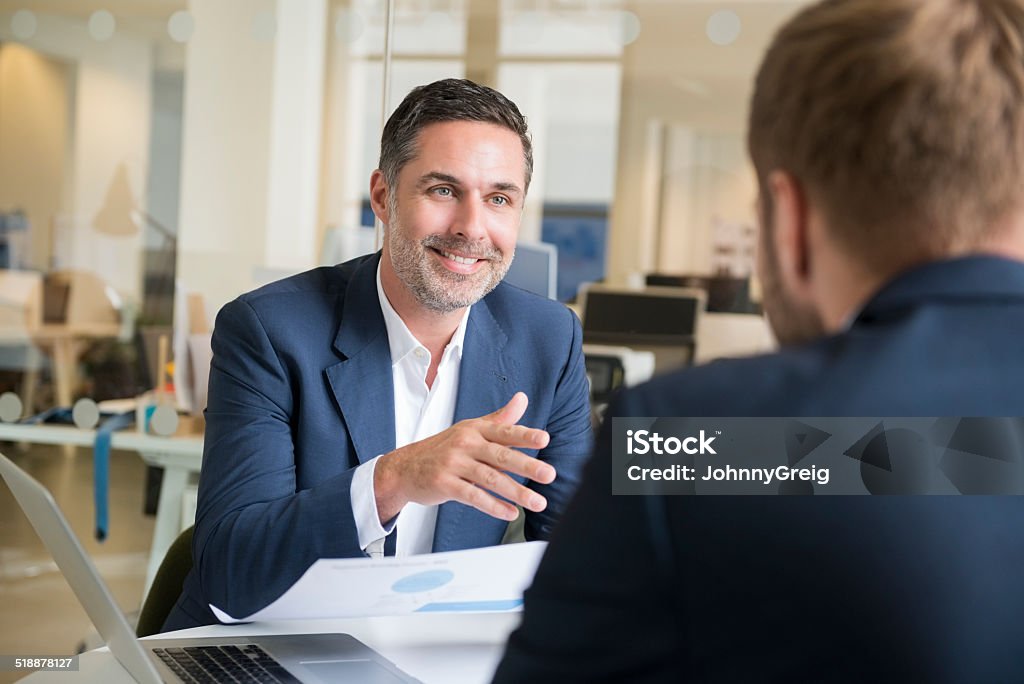 Businessman with colleague in meeting candid smiling businessman discussing with male colleague at desk in office Businessman Stock Photo
