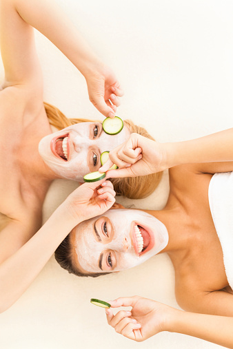Two female friends lying down on bed and wearing facial masks and cucumbers on eyes.