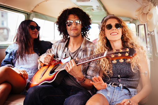 Two Multi Racial Hipsters, one girl and one man, playing guitar and singing together in the back of a van