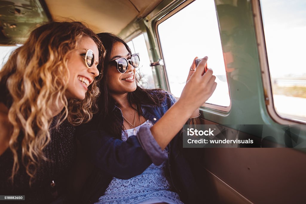 Two Hipster Girls Sitting Together taking a road trip photo Mixed race girl and caucasian blond friend smile while taking a road trip selfie in back of traveling retro van Teenage Girls Stock Photo