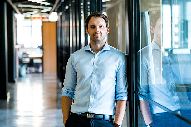 Confident businessman leaning on glass wall in office A photo of confident mid adult businessman standing with hands in pockets. Male executive is leaning on glass wall. He is in formals at office. man 30 years old stock pictures, royalty-free photos & images
