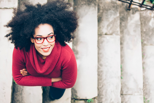 Portrait of smiling young woman standing arm crossed on staircase. High angle view. Woman with curly hair, red eyeglasses, and red pullover.