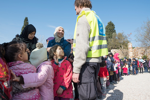Chios, Greece - March 18, 2016: Women and children refugees (arrived via Turkey) are standing in line waiting to being served a simple free breakfast (sandwich and water or juice) by international and local volunteers in a refugee camp on the Greek Island of Chios. A male volunteer from the Norwegian NGO Drop in the Ocean is communicating with a refugee mother.
