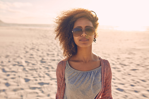 Portrait of attractive young woman standing on a beach. She is wearing sunglasses and looking at camera.