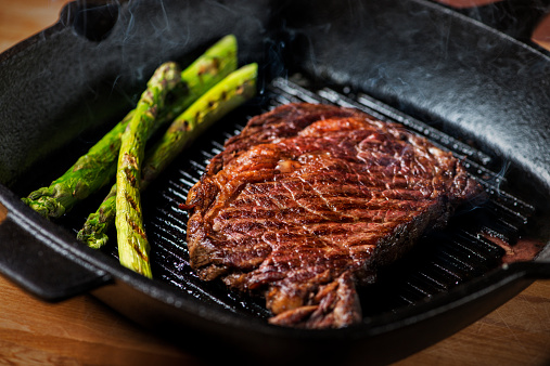 Ribeye steak grilled with asparagus, with visible smoke rising from the hot cast iron pan.  Shallow DOF.