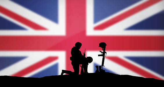A soldier kneeling in front of the grave of a fallen soldier. Great Britain flag in the background.