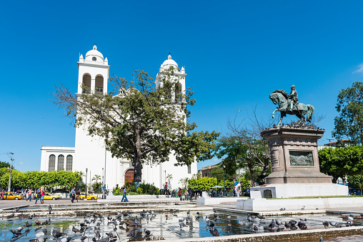 San Salvador, El Salvador - December 22, 2015: Church on la Ruta de las Flores in Las Salvador. Front view town square with memorial sculpture of Capitan General Gerardo Barrios 1909 and lots of pigeons in a puddle. In front of a churc some people and cars on the street. Clear blue sky in background. 