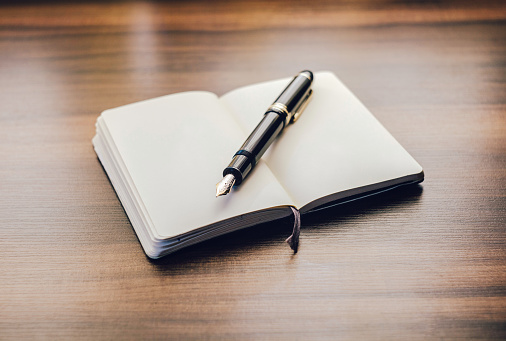 An open moleskin notebook with clean white pages and a black fountain pen with golden nib diagonally across over it. 