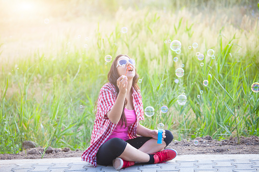 Young woman having fun outdoor. Sitting on the ground, legs crossed.and  blowing bubbles. Wear checkered shirt, leggins, red sneakers and sunglasses. Grassfield as background.