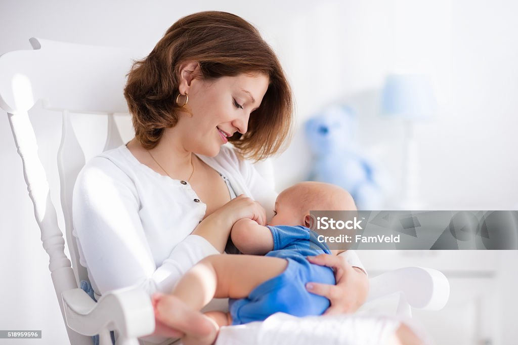 Nice young mother with baby boy at home Young mother holding her newborn child. Mom nursing baby. Woman and new born boy in white bedroom with rocking chair and blue crib. Nursery interior. Mother playing with laughing kid. Family at home Breastfeeding Stock Photo