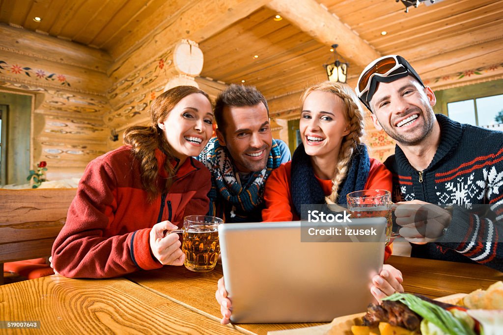 Friends after skiing Happy friends sitting in a mountain restaurant after skiing, enjoying beer and using a digital tablet together, smiling at the camera. Adult Stock Photo