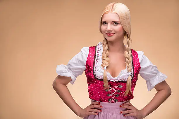 Young sexy smiling blonde wearing pink dirndl with white blouse looking at us putting her hands on her waist. Isolated on dark background
