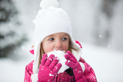 A six year old girl wearing a white hat, pink gloves and a winter jacket is eating a snowball. Snowflakes are falling against a blurred wintery background. Melted snow forms water drops on her face.