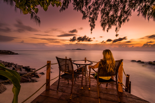 Rear view of a woman sitting on a chair at the beach and looking at breathtaking sunset.