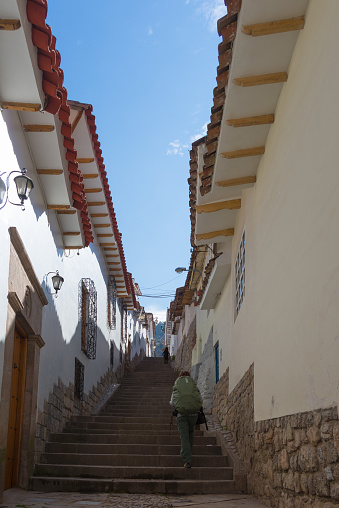 Backpacker walking uphill on staircase in a narrow alley of San Blas district, Cusco, Peru, former Inca capital, famous travel destination in the world. Wide angle view from below, morning bright light.