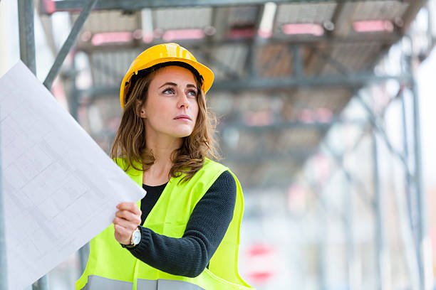 Young female engineer with office blueprints Female construction worker with helmet and safety jacket on construction site examining office blueprints. Outdoors foreman stock pictures, royalty-free photos & images