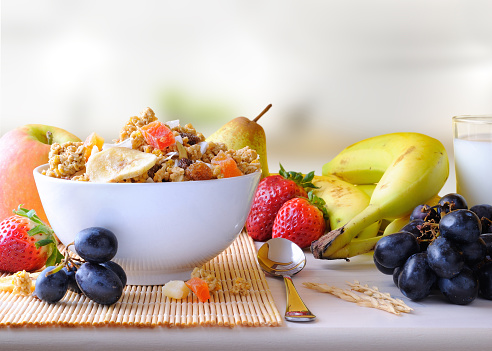 Bowl of cereal with fruit on a white wooden table and fresh fruits behind in the kitchen. Front view
