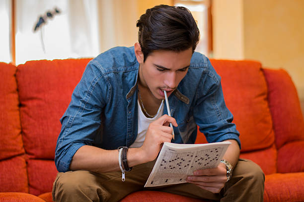 Young man sitting doing a crossword puzzle Young man sitting doing a crossword puzzle looking thoughtfully at a magazine with his pencil to his mouth as he tries to think of the answer to the clue crossword stock pictures, royalty-free photos & images