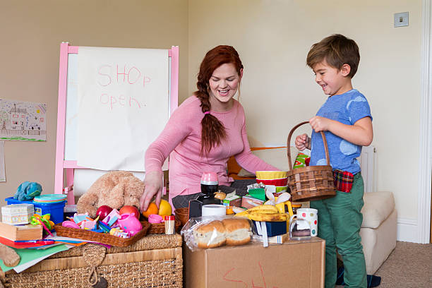 What shall I buy today? A pretend shop set up at home. Happy little boy holds a small basket and smiles. A woman sits surround by items for sale. Smiling she goes to pick up an item to sell the little boy. trans boy stock pictures, royalty-free photos & images