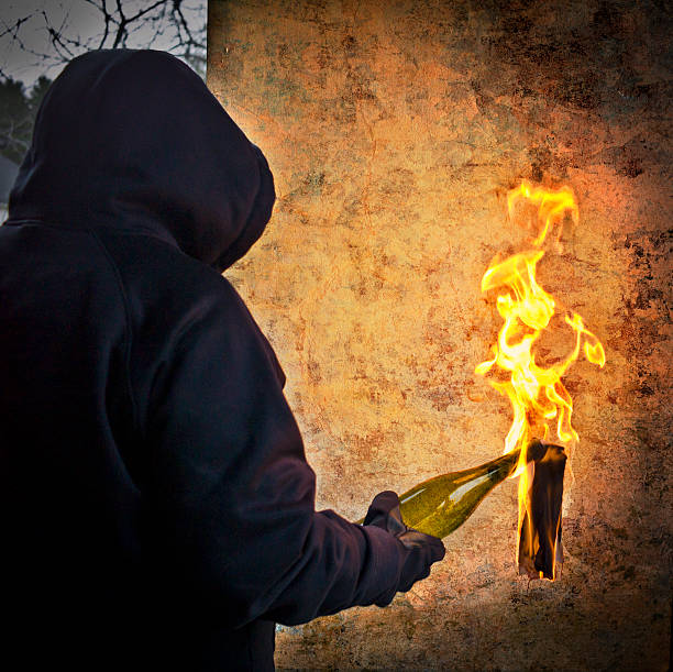 Hooded person holding a burning molotov cocktail.  Flames from the burning wick as they get ready to throw.