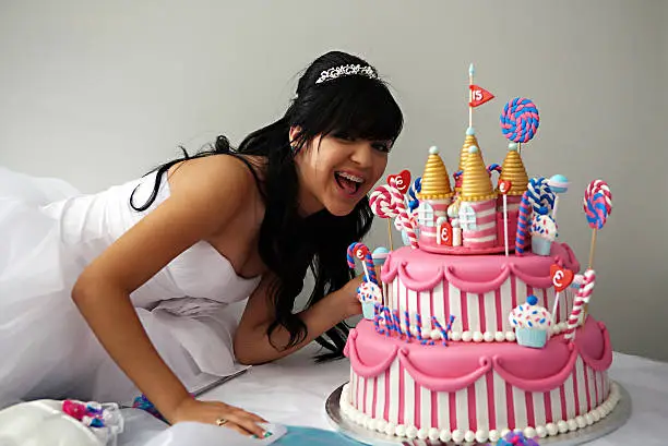 A 15 year old hispanic girl celebrates her birthday with a fancy birthday cake with a candyland theme. The three layer cake is covered with fondant and candy decorations.