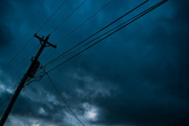 A leaning Telephone Pole with a transformer, carrying power lines, telephone lines and CATV cables with dark storm clouds moving in the background