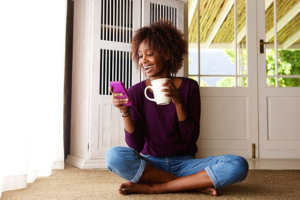 donna sorridente seduto sul pavimento a casa con il cellulare - sedere per terra foto e immagini stock