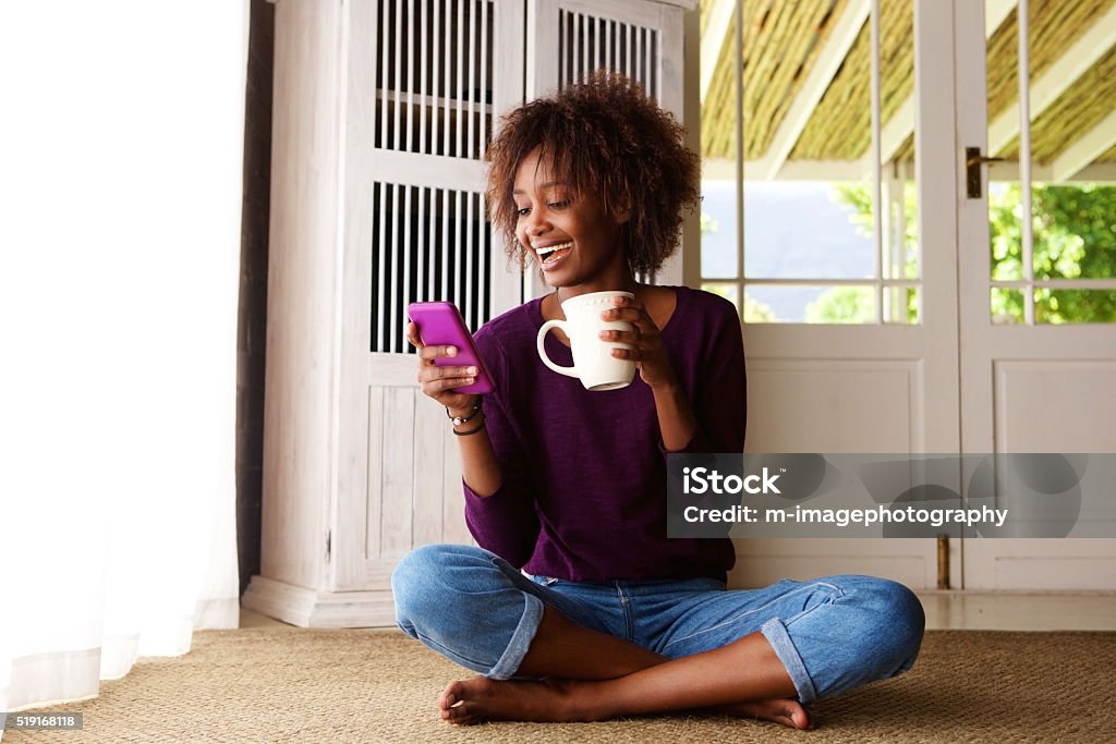Smiling woman sitting on floor at home with cell phone Portrait of a smiling young black woman sitting on floor at home with cell phone Women Stock Photo