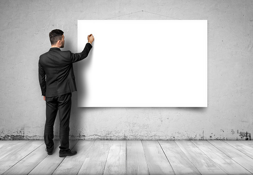 Back view of a businessman standing on a wooden floor and  drawing at a white empty canvas on a concrete wall