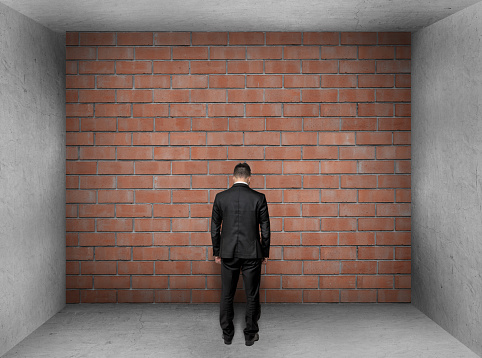 A businessman with bowed head stands in front of a brick wall in interior. Stalemate. Problem.