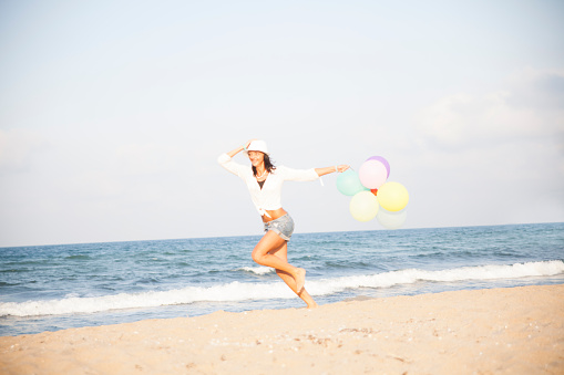 Young girl holding balloons and running on the beach. Wear white blouse and hat, short jeans, barefoot. Hand on hat. The sea and the waves as background..