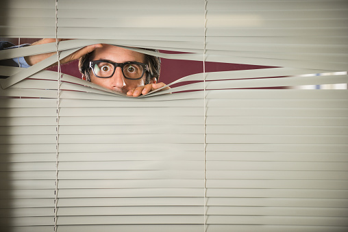 Nerdy startled man in glasses, mid thirties, staring through Venetian blinds.