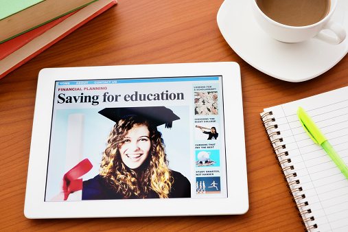 Photographer's images and design. The screen of a digital tablet shows a website with a pretty, smiling graduate and the headline: Saving for education. Books, a notebook, and a coffee cup also sit on the desk.