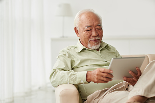 Senior Vietnamese man sitting on sofa and reading new on his tablet computer