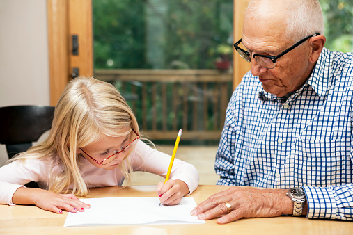 A 74 year old grandfather is helping his six year old granddaughter with her homework. She has drawn a line on a piece of paper and is counting using tally marks. The setting is a dining room table with patio door, deck and woods in the background.