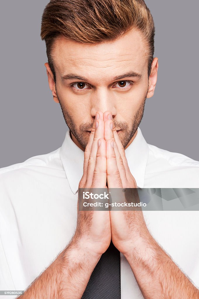 It is urgent question. Thoughtful young man in formalwear holding hands clasped near face while standing against grey background Adult Stock Photo