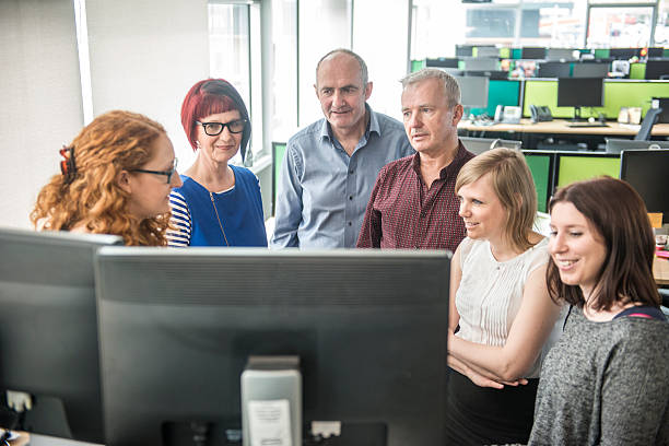 Creative team meeting at desk in office, all standing around two desktops, Dublin, Ireland. 