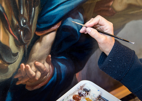 Restorer uses a thin brush to retouch pictorial imperfection on a painting surface.