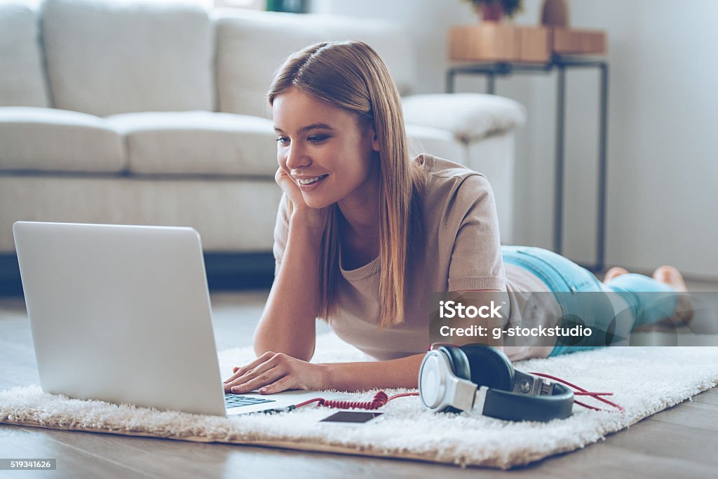 Relaxing at home. Beautiful young woman using her laptop while lying on carpet at home Laptop Stock Photo