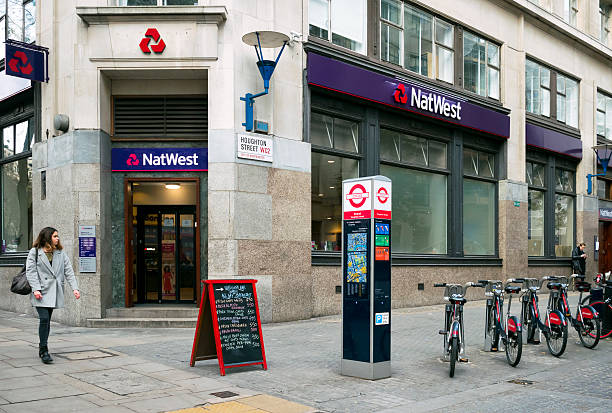 NatWest Bank and Santander bikes in Houghton Street, London London, England - November 23, 2015: A row of Santander ‘Boris Bikes’ in their dock outside a branch of NatWest bank on the corner of Houghton Street and Aldwych, Central London. A woman is looking up Houghton Street and a man is studying his phone. A handwritten sign points to a nearby cafe bar. (Overcast day.) bicycle docking station stock pictures, royalty-free photos & images