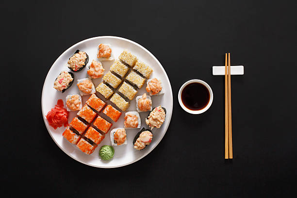 Set of sushi maki and rolls at black background Japanese food restaurant, sushi maki gunkan roll plate or platter set. Chopsticks, ginger and wasabi. Sushi at white round plate, black background. Top view with soy sauce sushi round plate stock pictures, royalty-free photos & images