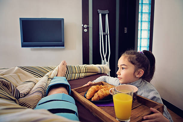 Knee Injury /ACL,PCL/ Little girl is helping her father who is lying in the bed after knee surgery bringing him breakfast in bed. posterior cruciate ligament stock pictures, royalty-free photos & images