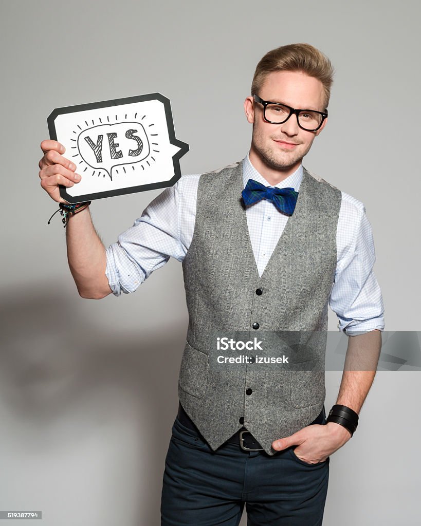 Fashionable blonde young man wearing tweed vest and bow tie Portrait of confident blonde young man wearing tweed vest, bow tie and nerd glasses, holding speech bubble with word "yes" in hand, smiling at camera. Studio shot, grey backgound, one person.  Adult Stock Photo