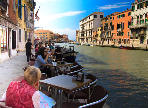 Venice, Italy - May 16, 2015: Diners and tourists sitting at a restaurant-cafe at the edge of a canal in the Cannareggio neighborhood of Venice, Italy. The tourist in the foreground is studying a map of Venice.