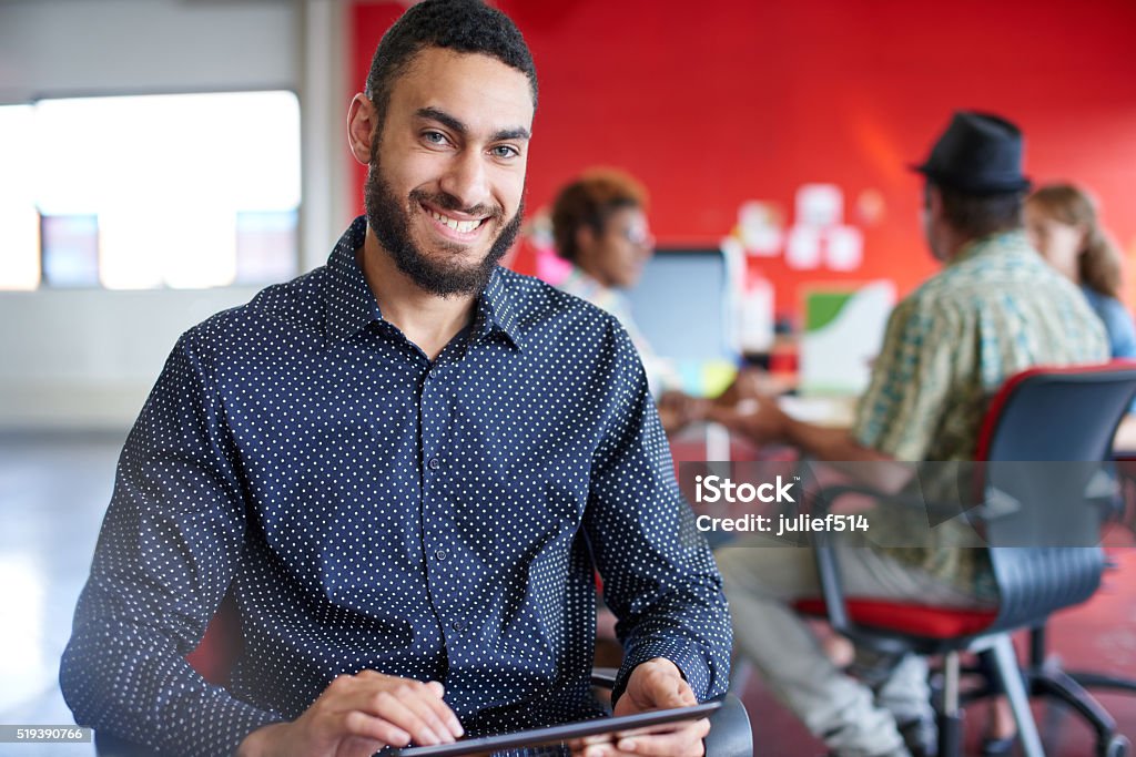 Confident male designer working on a digital tablet in red Casual portrait of a business man using technology in a bright and sunny startup with the team in the background Red Stock Photo