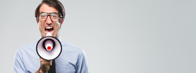 Nerdy caucasian businessman mid thirties, wearing a blue shirt and tie, angrily yelling into megaphone. Shot in the studio on a grey background on the Nikon D800.