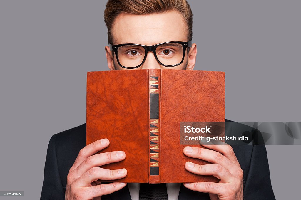 More knowledge! Young man in formalwear hiding his face behind a book while standing against grey background Adult Stock Photo